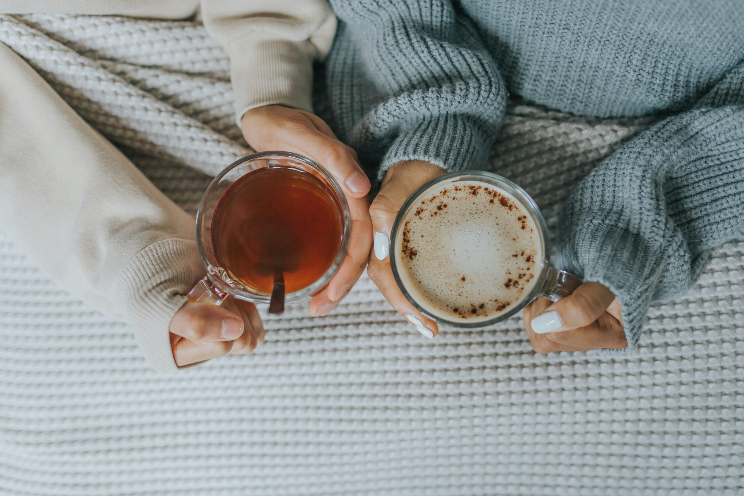 Cozy overhead view of a woman’s hands holding a warm drink while resting on soft neutral bedding, capturing a calm, low‑energy morning moment.