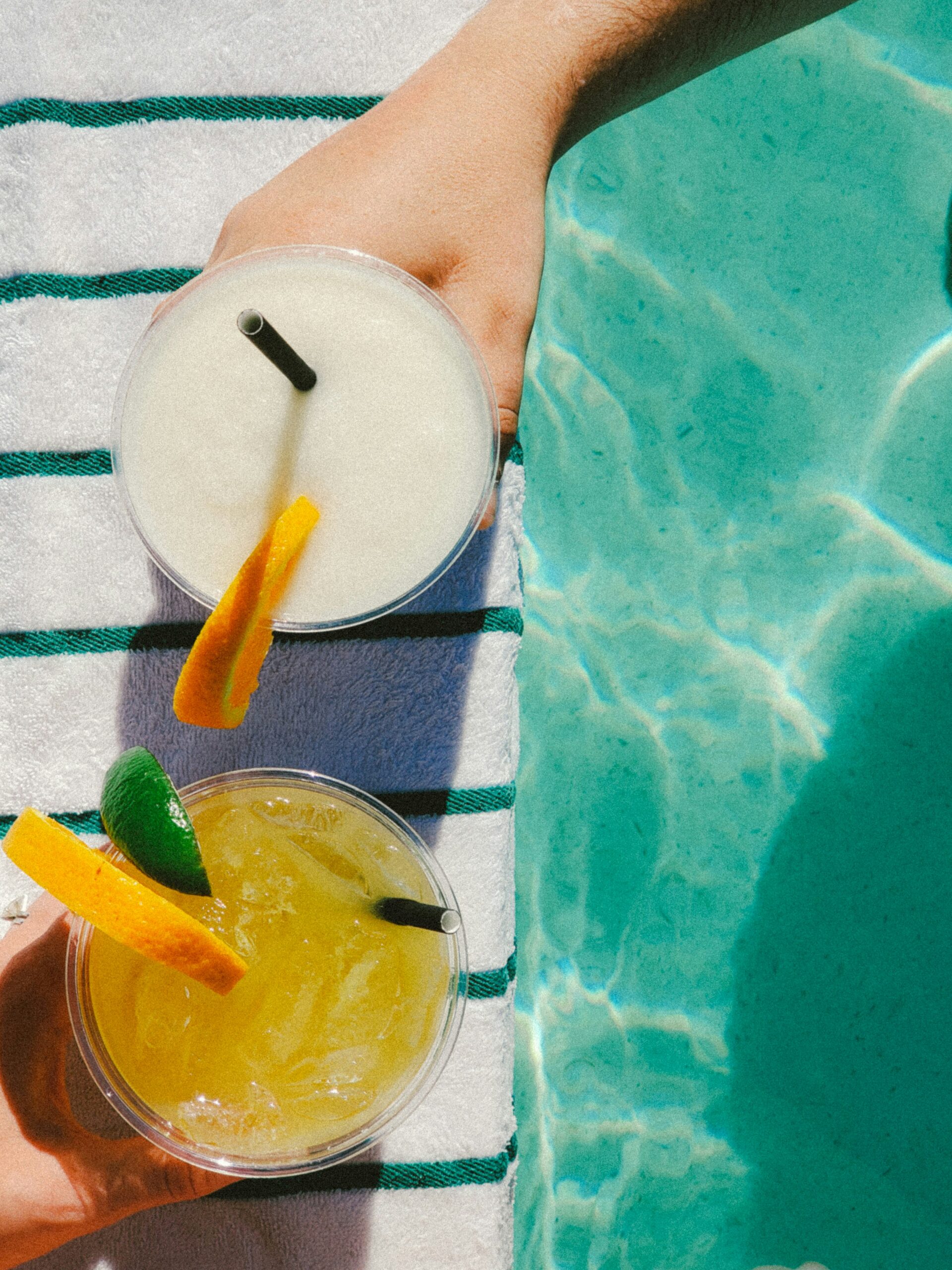 Two hands holding refreshing summer drinks beside a swimming pool, one a frozen white drink with an orange slice and the other a yellow iced drink with citrus garnishes, resting on a striped towel in bright sunlight.