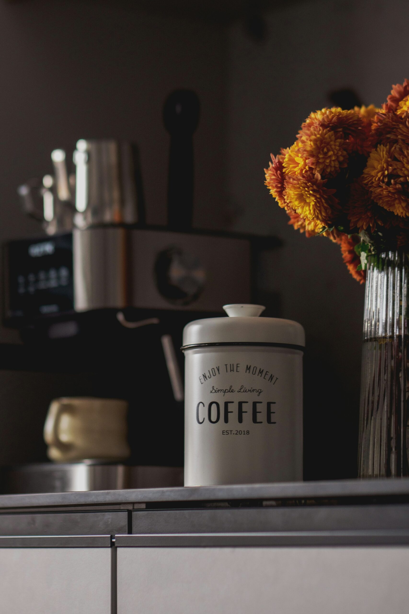 A warm, softly lit kitchen counter with a white coffee canister, fresh orange and yellow flowers in a glass vase, and a gold mug beside an espresso machine, creating a calm and cozy evening atmosphere.